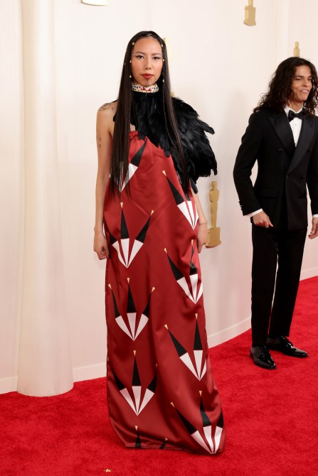 Quannah Chasinghorse at the 2024 Oscars red carpet in a red printed gown with black feather accents