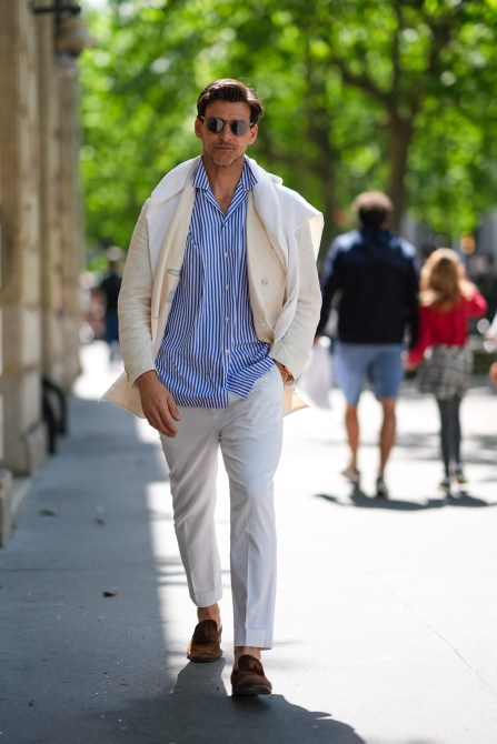 Man wears black sunglasses, a white and blue striped button up long sleeve shirt, cream blazer jacket, a white sweater around shoulders, white straight fitted pants, dark brown loafer moccasin suede shoes,