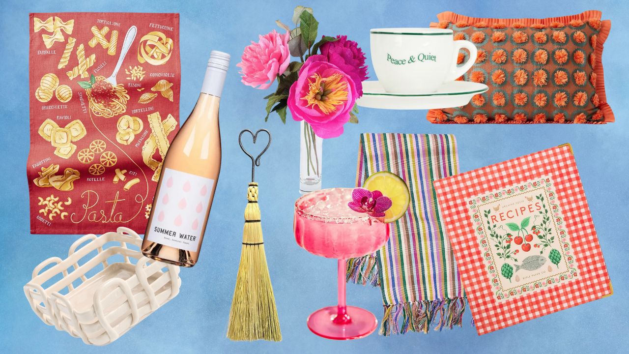 From left to right: A red pasta towel, a white ceramic basket, a bottle of wine, pink paper flowers, a broom, a pink coupe glass, a stripe towel, a red recipe book, a white and green espresso cup, an orange and blue pillow