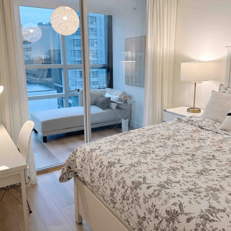 A bedroom with part of a big bed, a desk, a nightstand, a lamp, and an open den in the back. A downtown Vancouver Airbnb.