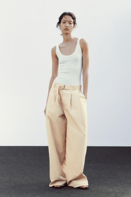 A model wearing the Zara Pleated Belted Pants in Vanilla colorway with a plain white tank top