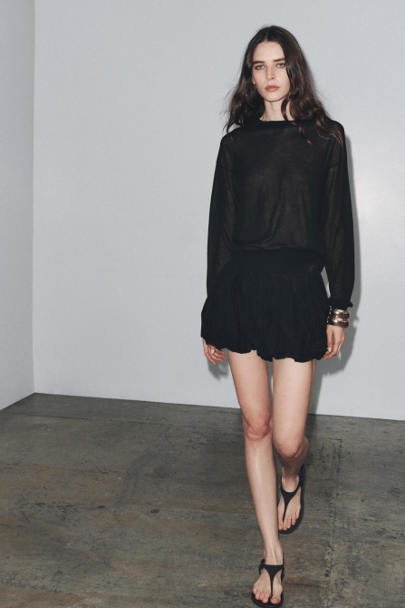 A model wearing the Zara Balloon Skort in black with a black long-sleeve shirt and black thong sandals and chunky bracelets